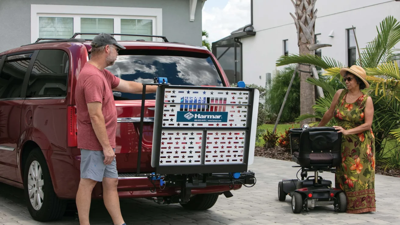 A man secures a mobility scooter on a rear-mounted carrier of a red van, while a woman in a floral dress and hat stands nearby on a driveway.