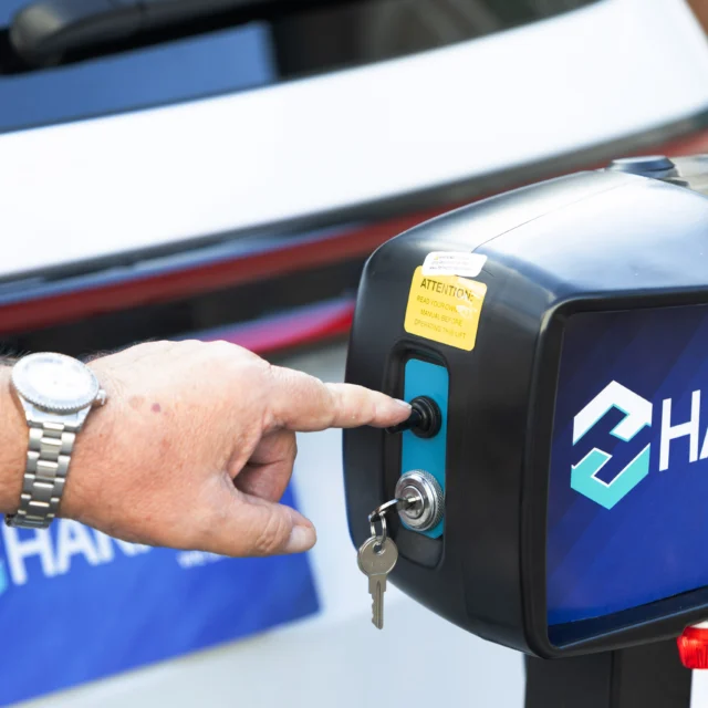 Person pressing a button on a device labeled "HARMAR," with a key inserted below. A watch is visible on the person's wrist.