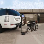 A wheelchair lift attached to the rear of a white SUV near a building on a clear day.