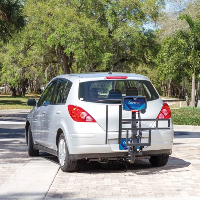 A silver car with a mobility scooter carrier attached to the rear, parked on a paved road surrounded by trees and greenery.