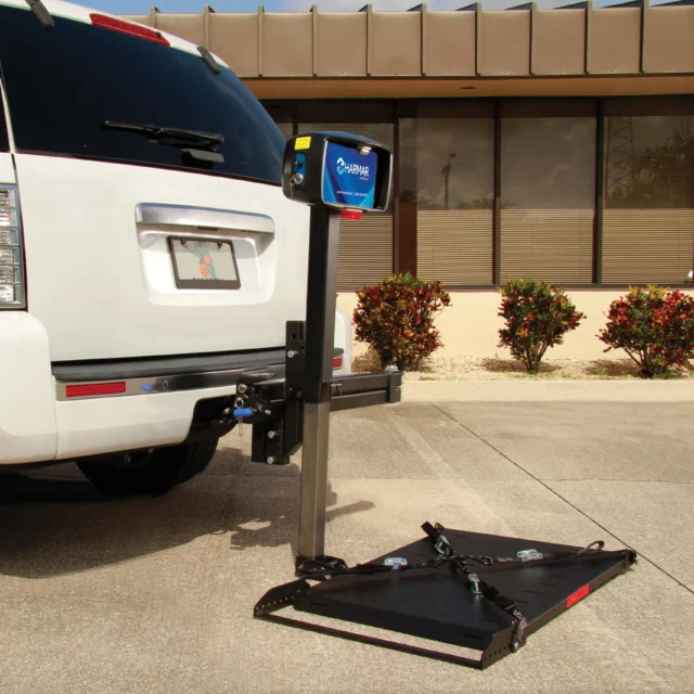 White SUV with a mobility lift attached to the rear, parked in a paved area near a building with bushes.
