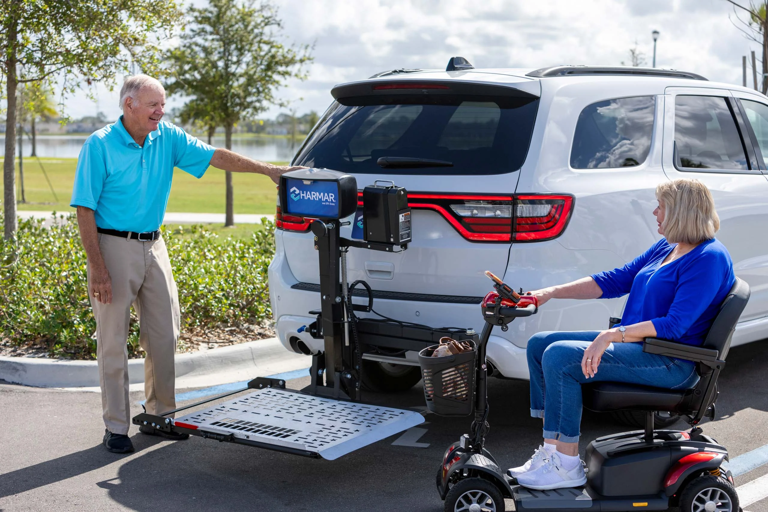 An elderly man and woman use a vehicle lift to load a mobility scooter onto the back of a white SUV in a parking lot.