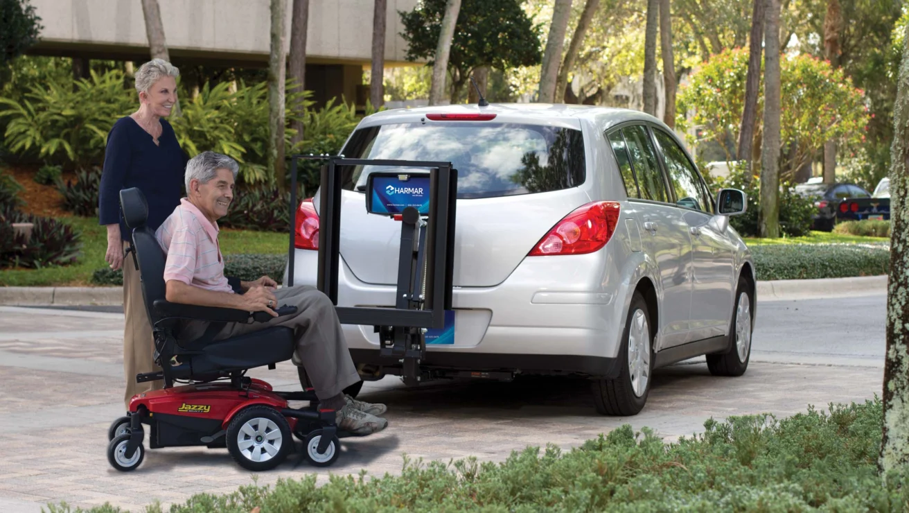 A person in a motorized wheelchair uses a lift attached to the back of a parked hatchback car, with another individual observing. Trees and a building are in the background.