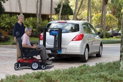 A person in a motorized wheelchair uses a lift attached to the back of a parked hatchback car, with another individual observing. Trees and a building are in the background.