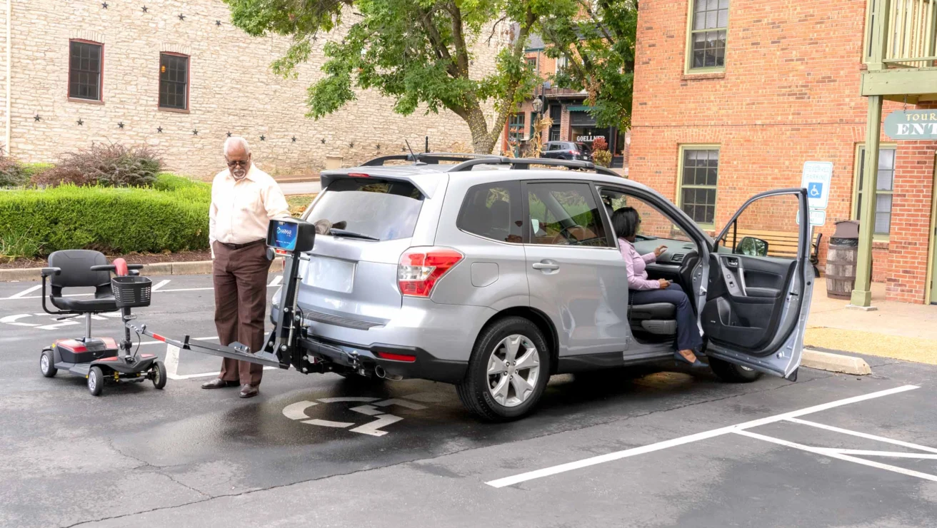 Man standing by an SUV with a mobility scooter attached to the back. Another person is seated inside the vehicle, which is parked in a designated parking area.