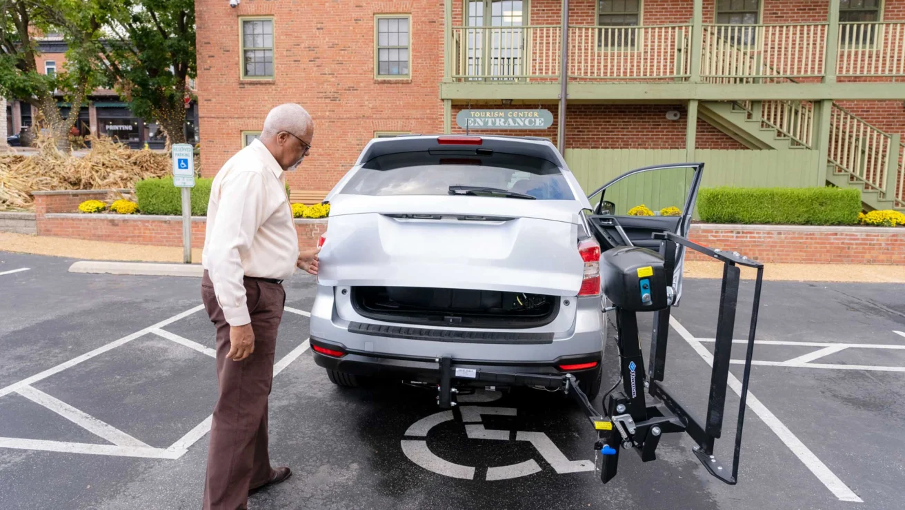 An elderly man stands by an SUV in a parking spot marked for disabilities. The SUV has a wheelchair lift attached to its rear.