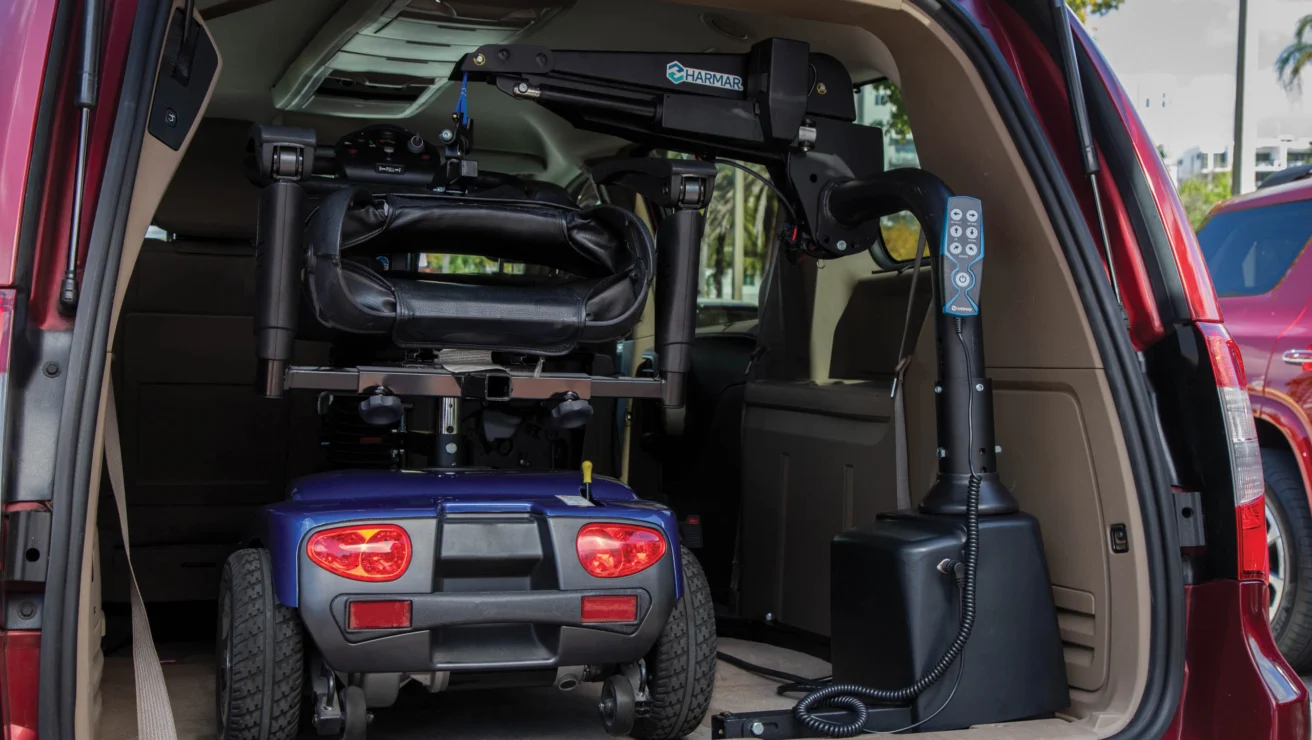 A red vehicle with an open trunk shows a motorized wheelchair secured on a lift mechanism for transport.