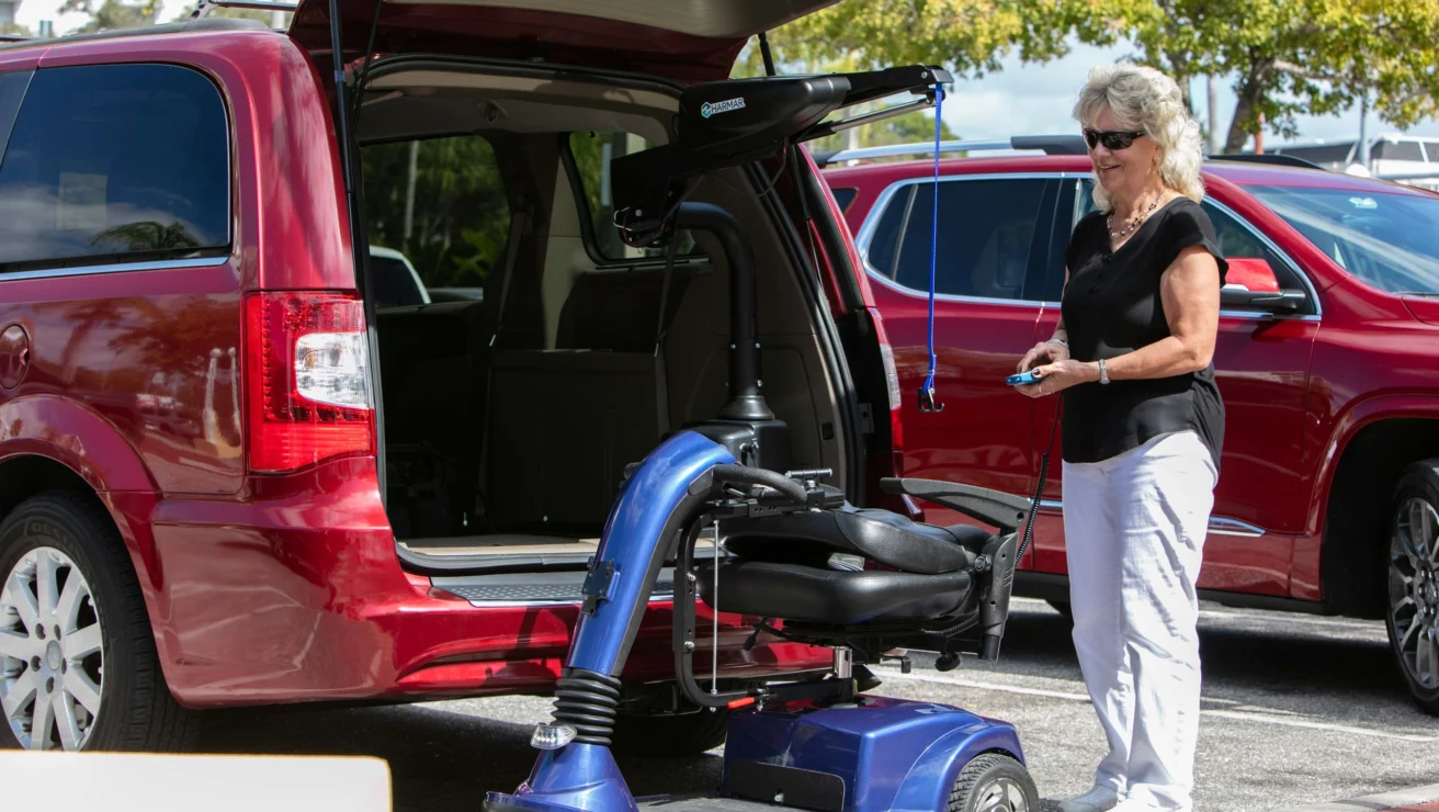 Woman using a motorized carrier lifts a blue mobility scooter into the back of a red minivan in a parking lot.