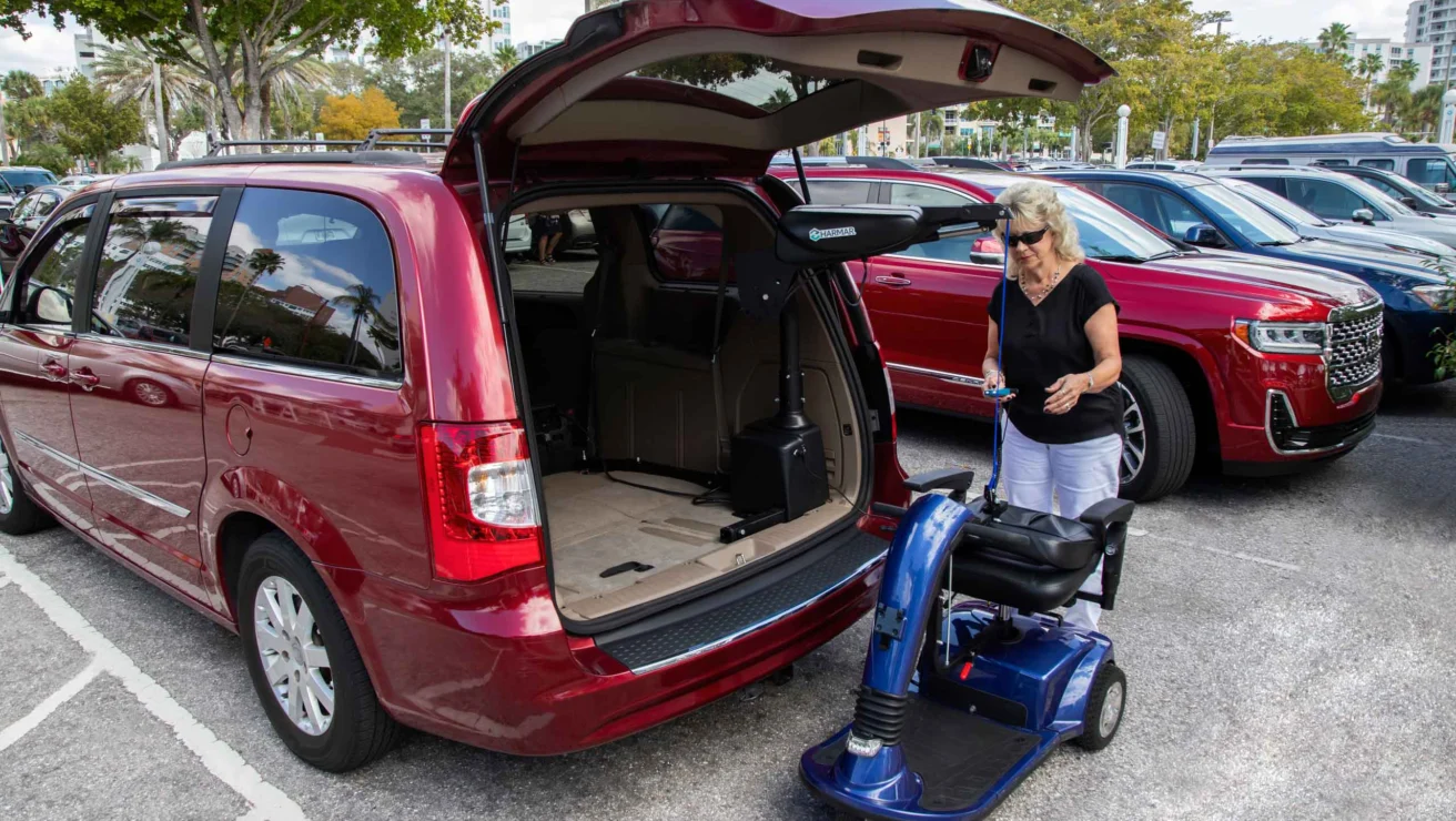 A woman places an electric mobility scooter into the back of a red minivan in a parking lot.