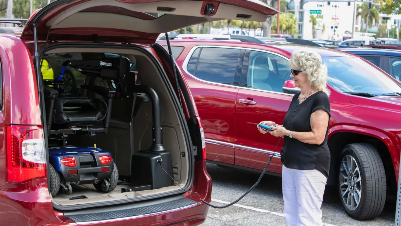 An older woman operates a lift to load a mobility scooter into the back of a red SUV in a parking lot.