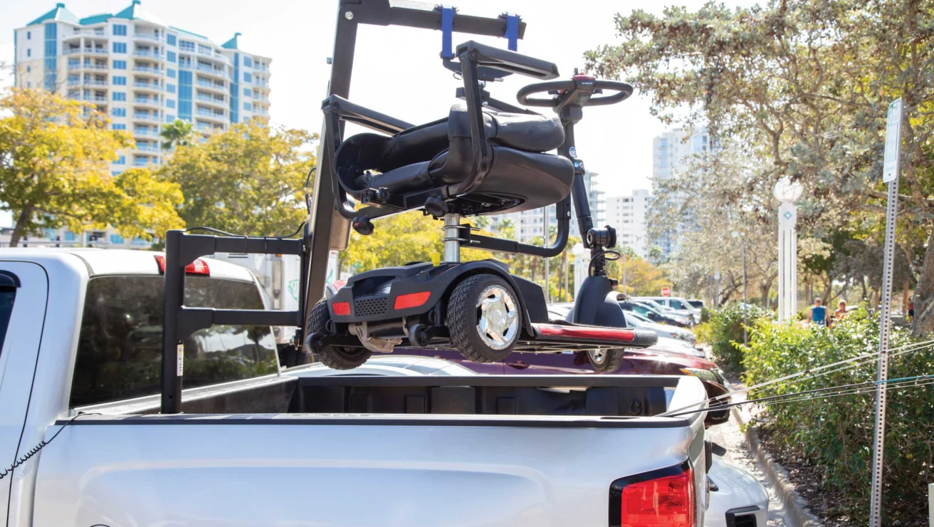 Motorized scooter mounted on a lift attached to a white pickup truck in a parking lot, with buildings and trees in the background.