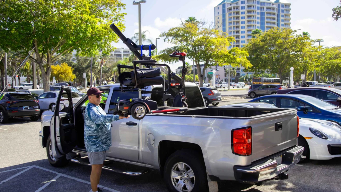 A man stands next to a silver pickup truck with an open tailgate. He is adjusting a motorized wheelchair stored in the truck bed. The parking lot has several cars and nearby tall buildings.