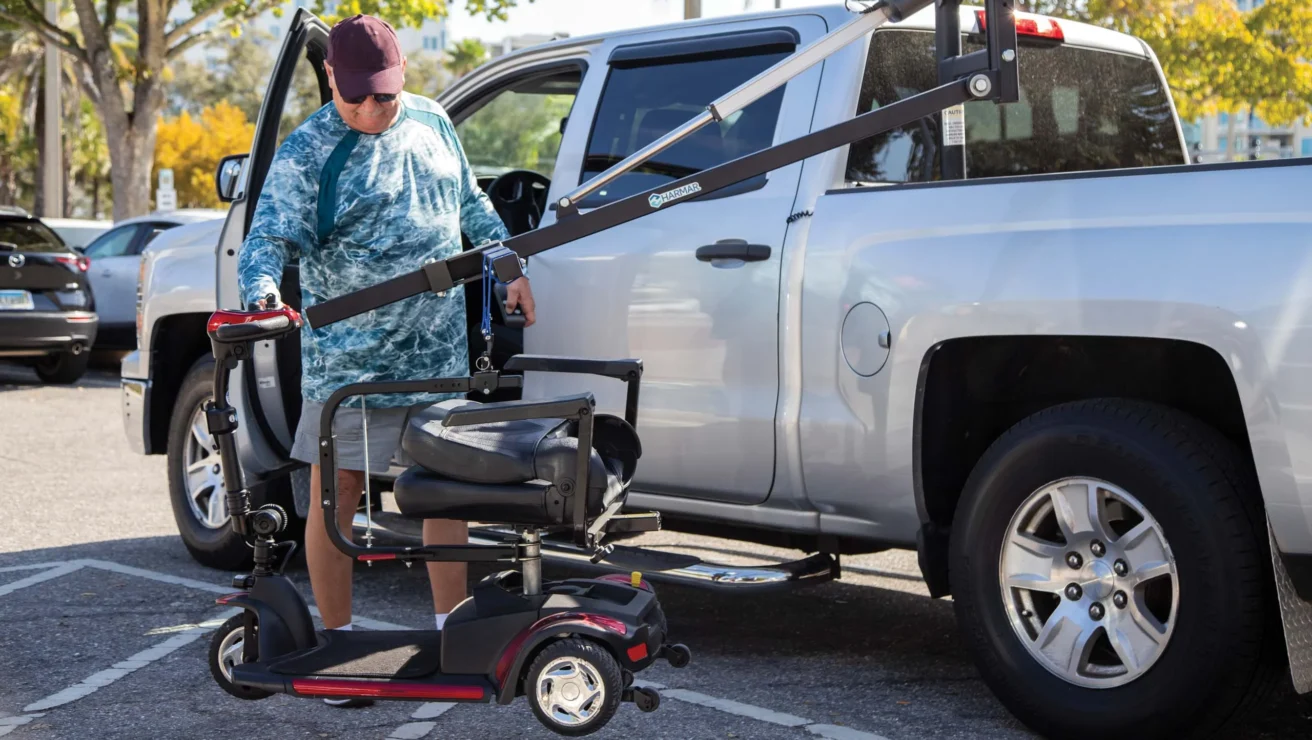 A person uses a lift to load a mobility scooter into the back of a silver pickup truck in a parking lot, surrounded by trees and other parked cars.