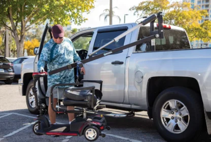 A person uses a lift to load a mobility scooter into the back of a silver pickup truck in a parking lot, surrounded by trees and other parked cars.