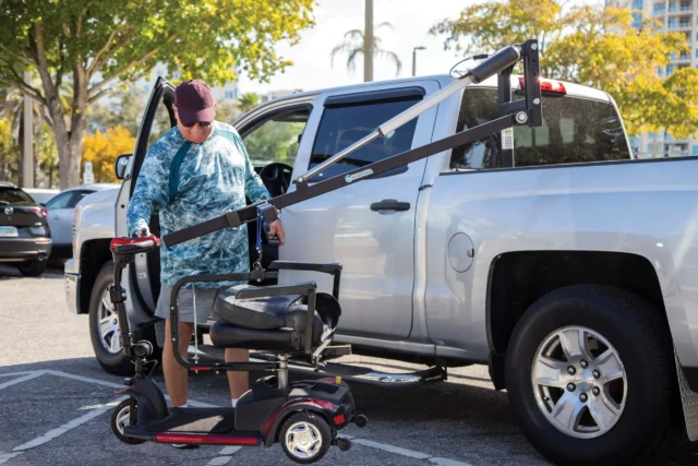A person uses a lift to load a mobility scooter into the back of a silver pickup truck in a parking lot, surrounded by trees and other parked cars.