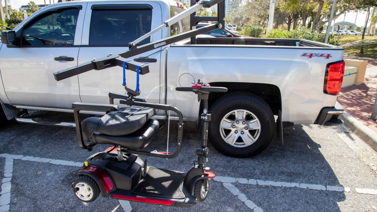 A mobility scooter is being lifted by a crane attached to the back of a silver pickup truck parked in a lot.