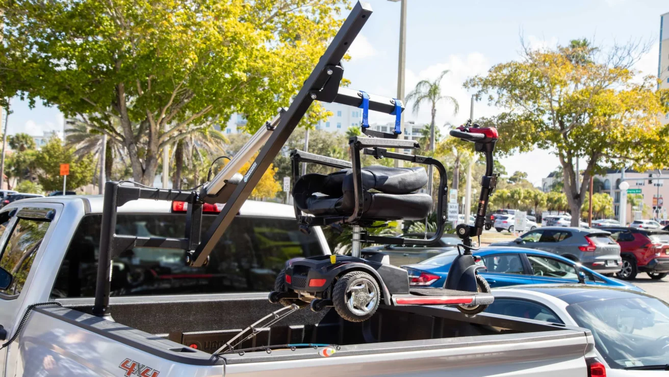 An electric wheelchair is secured on a lift attached to the bed of a white pickup truck in a parking lot.