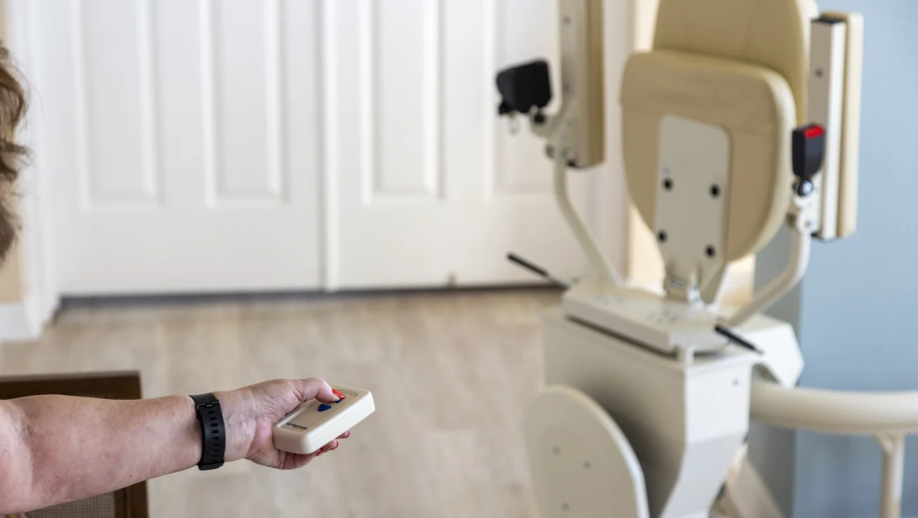 A person holds a remote control aimed at a beige stair lift positioned at the bottom of a staircase.