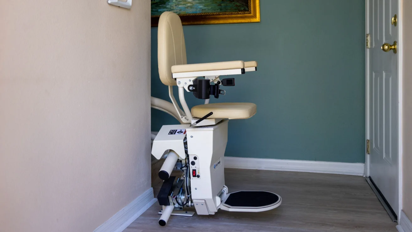 A stairlift is installed on a staircase, featuring a padded seat with armrests and a footrest, situated beside a wall and a painting in a residential setting.