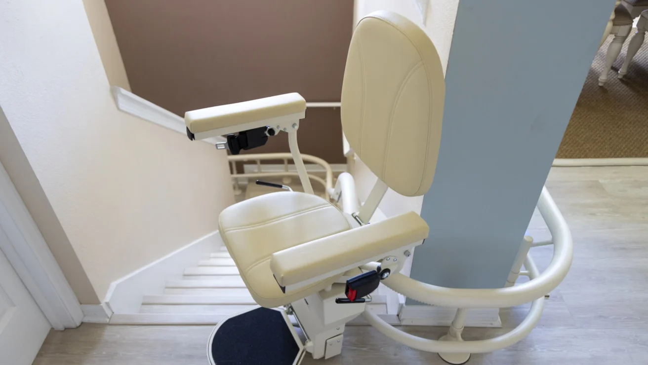 A beige stairlift is installed on a staircase, featuring a padded seat, armrests, and a footrest.