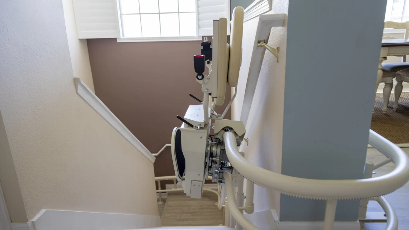A stairlift is installed along a residential staircase, with the chair folded up against the rail. A table and chairs are visible in the background.