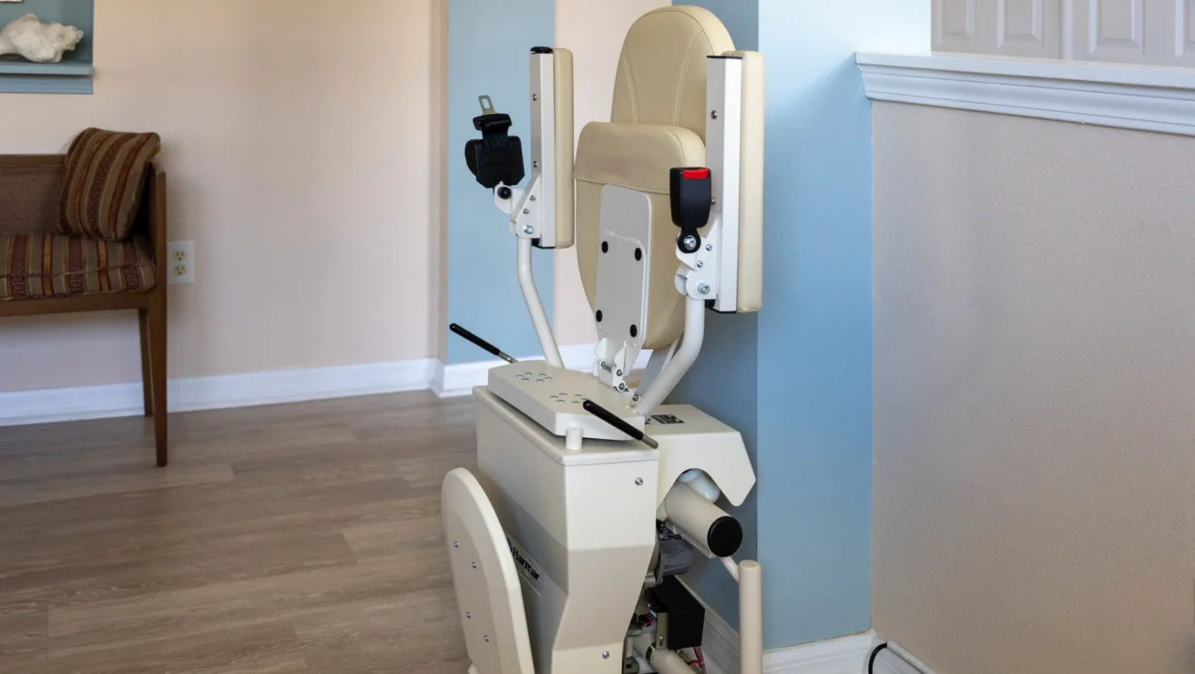 A stair lift is installed along the wall in a home, near a chair. The lift is folded up in its resting position on the wooden floor.