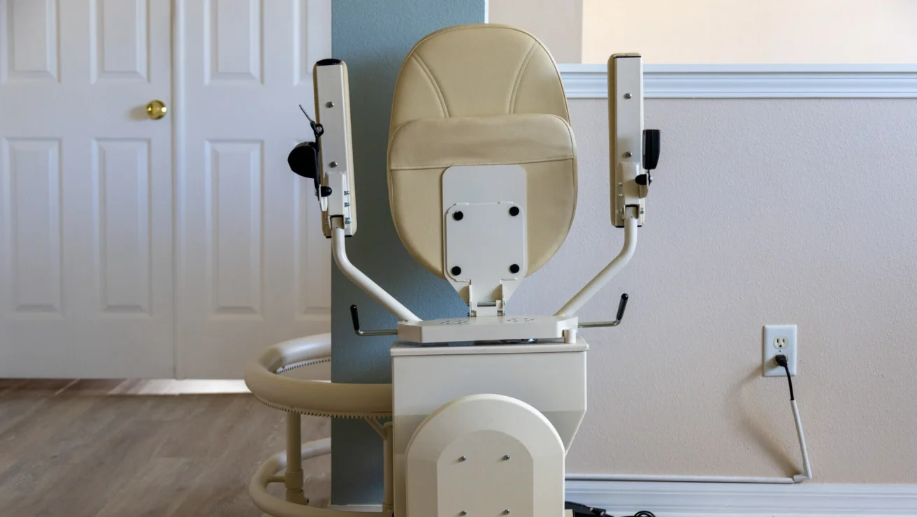 A stairlift chair positioned along a staircase, with beige upholstery and a mechanical track, in a residential setting.