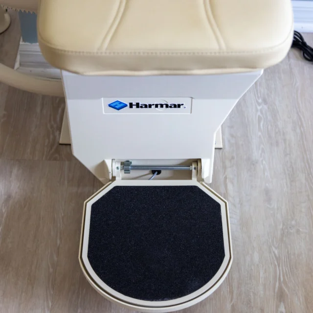 Stairlift footrest with a beige seat, labeled "Harmar," on a wooden floor.