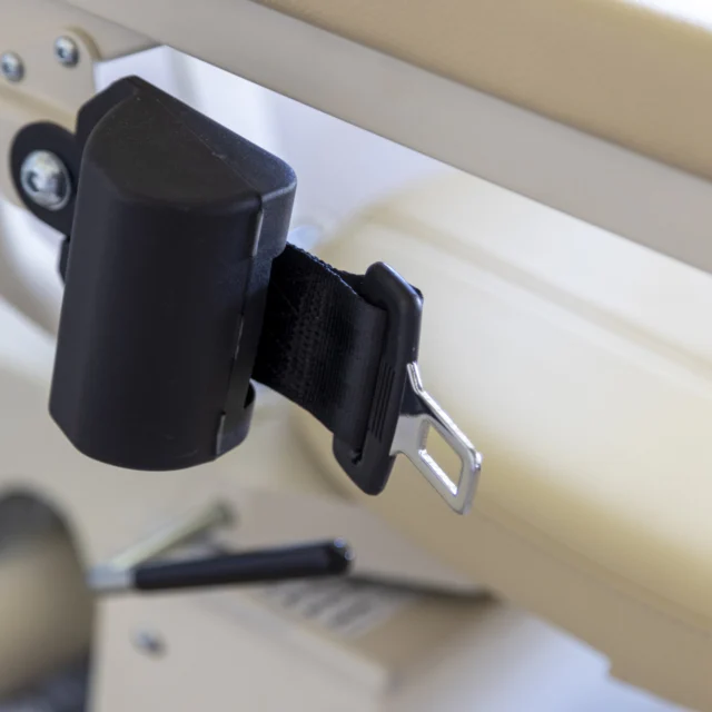 Close-up of a black seat belt buckle on a tan seat, with stitching detail and a lever visible.