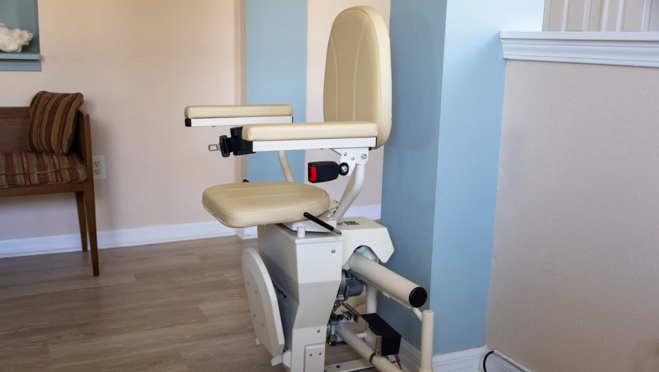 A beige stairlift is installed along a staircase. It has a padded seat, armrests, a seatbelt, and a footrest. The lift is positioned at the bottom of the stairs.