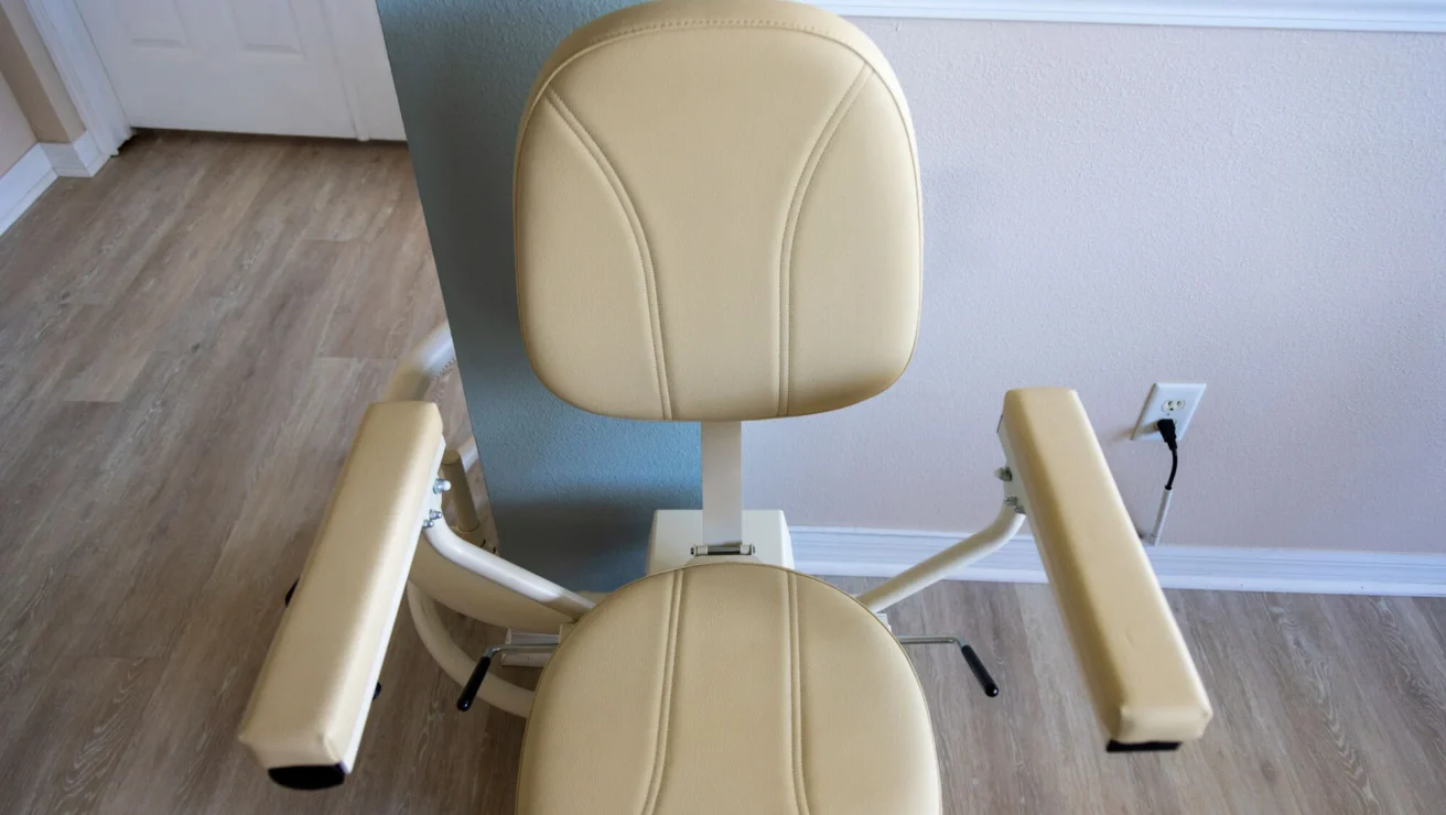 Beige stair lift chair with armrests and backrest, installed on a curved rail against a wall.