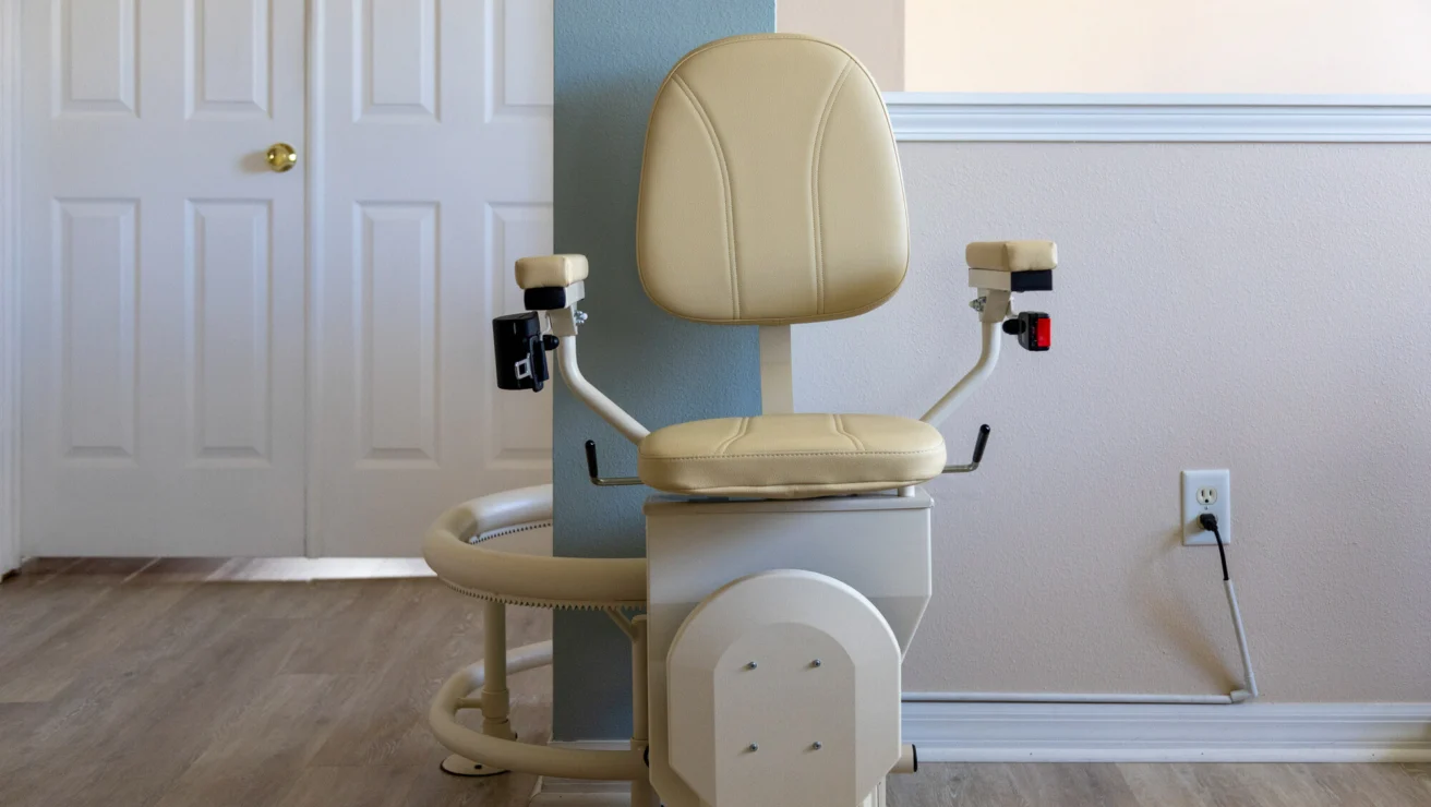 A beige stairlift with a cushioned seat and armrests is installed on a staircase beside a light blue wall in a residential setting.