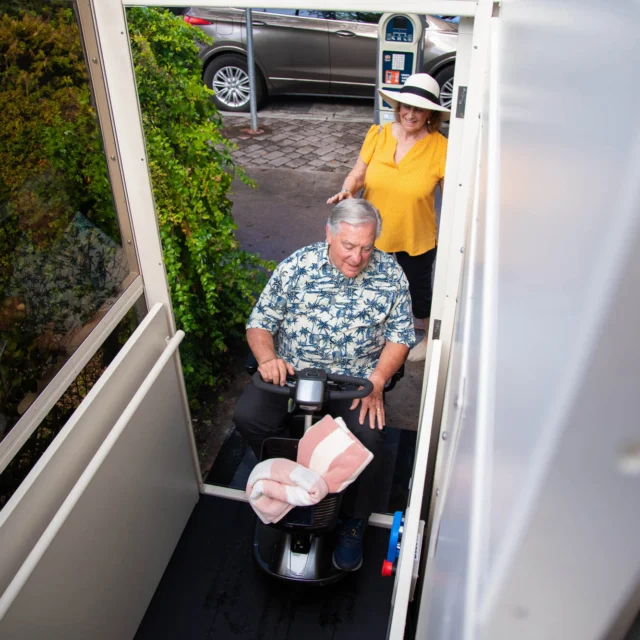 A person on a mobility scooter using a home lift with another person standing behind, outside a house with greenery and a parked car visible.