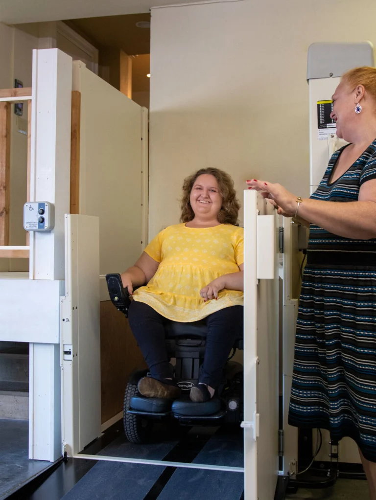 A woman in a wheelchair is on a platform lift. Another woman stands beside, smiling. They are in a garage with a wooden railing and a doorway in the background.