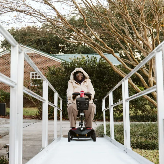 Person in a hoodie using a mobility scooter on a ramp leading to a red brick building, surrounded by trees and garden lights.