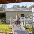A woman in a wheelchair descends a ramp outside a house, under a covered area. Trees and a fence are visible in the background.
