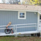 A person in a wheelchair ascends a white access ramp leading to the entrance of a light blue house with a brown roof.