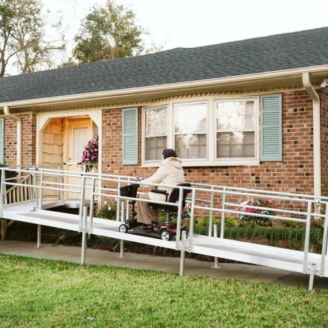 A person using a motorized wheelchair ascends a metal ramp leading to the entrance of a brick house with light blue shutters.
