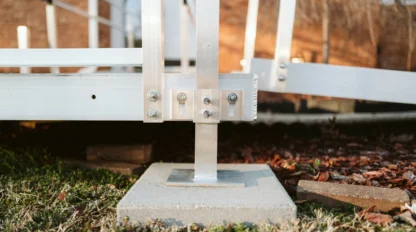 Metal support structure on a concrete base with bolts and nuts, surrounded by grass and mulch, near a brick wall.