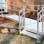 Aluminum wheelchair ramp with railings leading to a door on a brick house, placed over steps and grass.