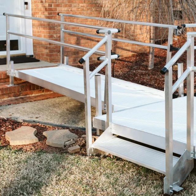 Aluminum wheelchair ramp with railings leading to a door on a brick house, placed over steps and grass.