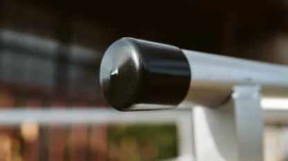 Close-up of a metal handrail with a black end cap, set against a blurred background of a building and greenery.