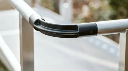 Close-up of a metal handrail with black connectors, part of a wheelchair ramp. The background shows blurred greenery and part of the ramp.