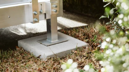 Close-up of a metal support beam anchored to a concrete slab on grass, surrounded by small plants.
