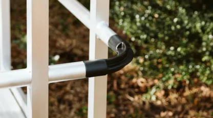 Close-up of an aluminum handrail with black connectors on a ramp, set against a brick wall and green shrubs.