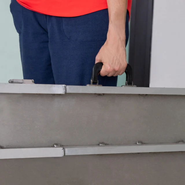 A person in a red shirt and blue pants holds the handle of a large metal case.