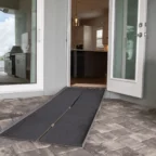 A foldable ramp placed on brick pavers leads to an open glass door of a house, enhancing wheelchair accessibility into the kitchen area.