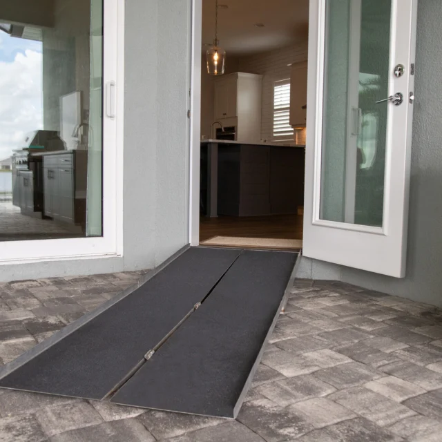 A foldable ramp placed on brick pavers leads to an open glass door of a house, enhancing wheelchair accessibility into the kitchen area.