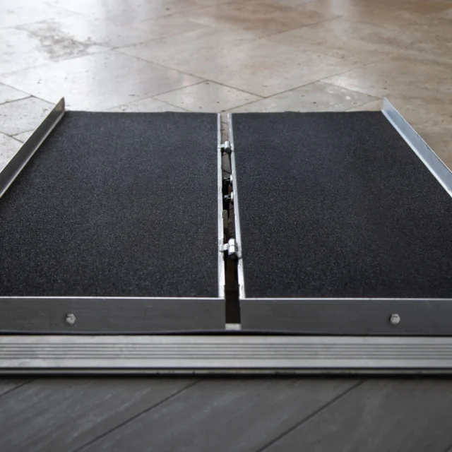 Foldable wheelchair ramp placed at an entrance on tile flooring, featuring a non-slip surface and metallic edges.