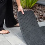 Person in sandals adjusting a textured black rubber mat on a paved surface outdoors.